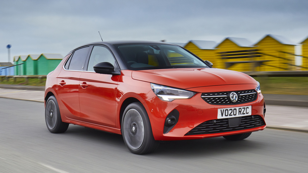 A fiery orange Vauxhall Corsa cruises down a seafront road next to brightly coloured beach huts